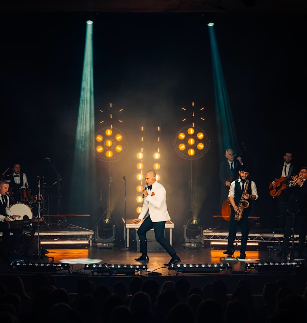 Luke Thomas in a white tuxedo jacket dancing centre stage with the band behind him under warm stage lights and teal spotlights, in front of a packed theatre